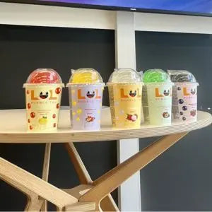 Five colourful LOL Bubble Tea cups with a brand logo on a wooden table against a blackboard background